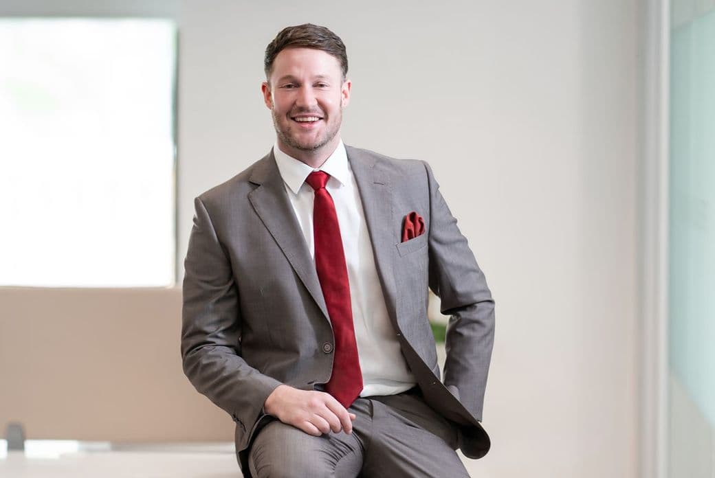 Smiling man in a gray suit with a red tie and pocket square, seated on a desk in a bright office.