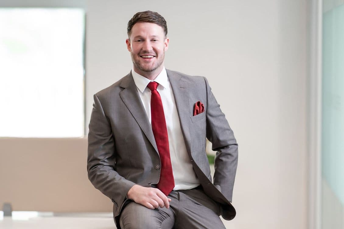 Smiling man in a gray suit with a red tie and pocket square, seated on a desk in a bright office.