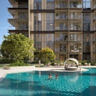 Modern apartment building with glass balconies, trees, and a pool. People are swimming and relaxing around the pool on a sunny day.