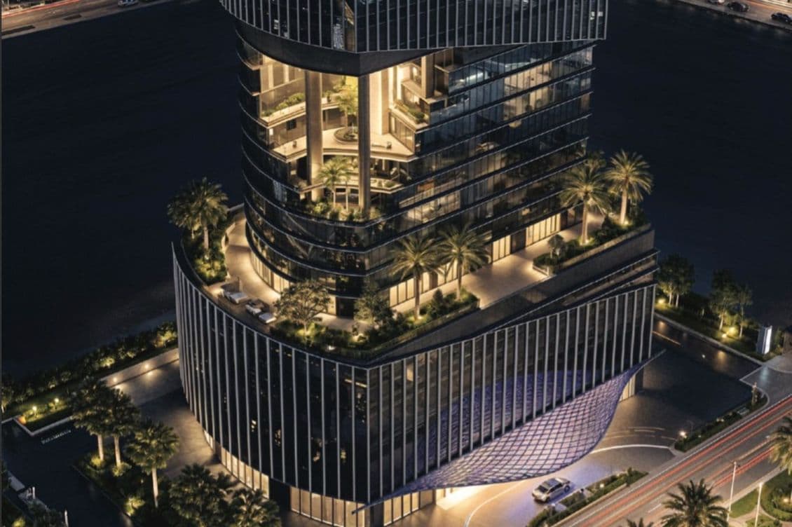Aerial view of a modern, illuminated multi-story building with glass facade, palm trees, and urban surroundings at night.