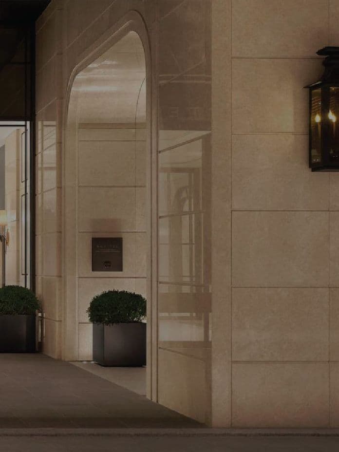 Elegant hotel entrance with glass doors, potted plants, and a lit wall sconce on a stone facade.
