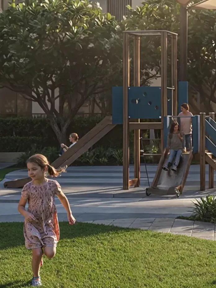 Children playing in a sunlit playground, with two kids running on grass and others using a slide, surrounded by trees and parents watching nearby.