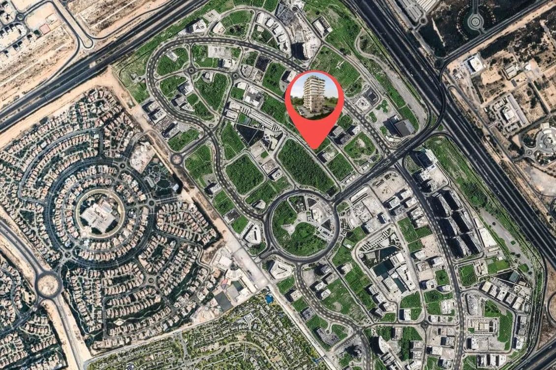 Aerial view of a cityscape with green parks and circular roads. A red map pin marks a specific building within the area.