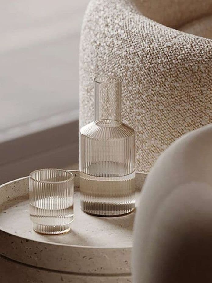 Textured glass carafe and tumbler on a round stone table beside a light beige, textured armchair in a minimalist setting.