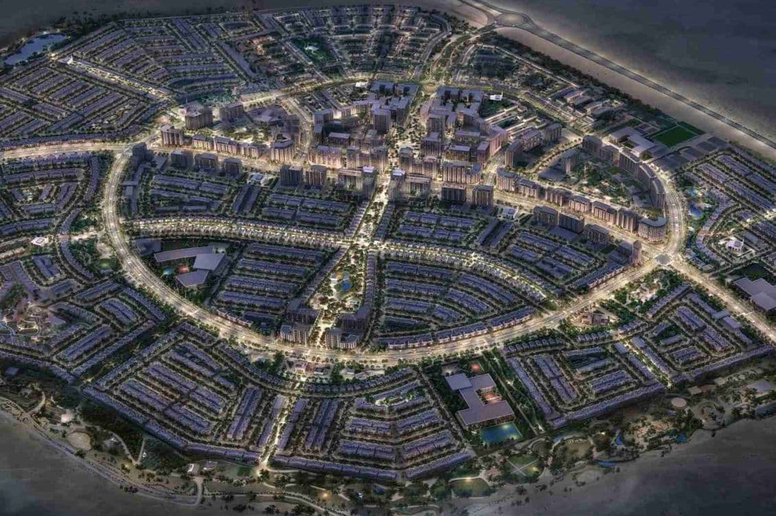 Aerial night view of a planned city with illuminated streets forming concentric circles, surrounded by a grid of residential areas near a coastline.