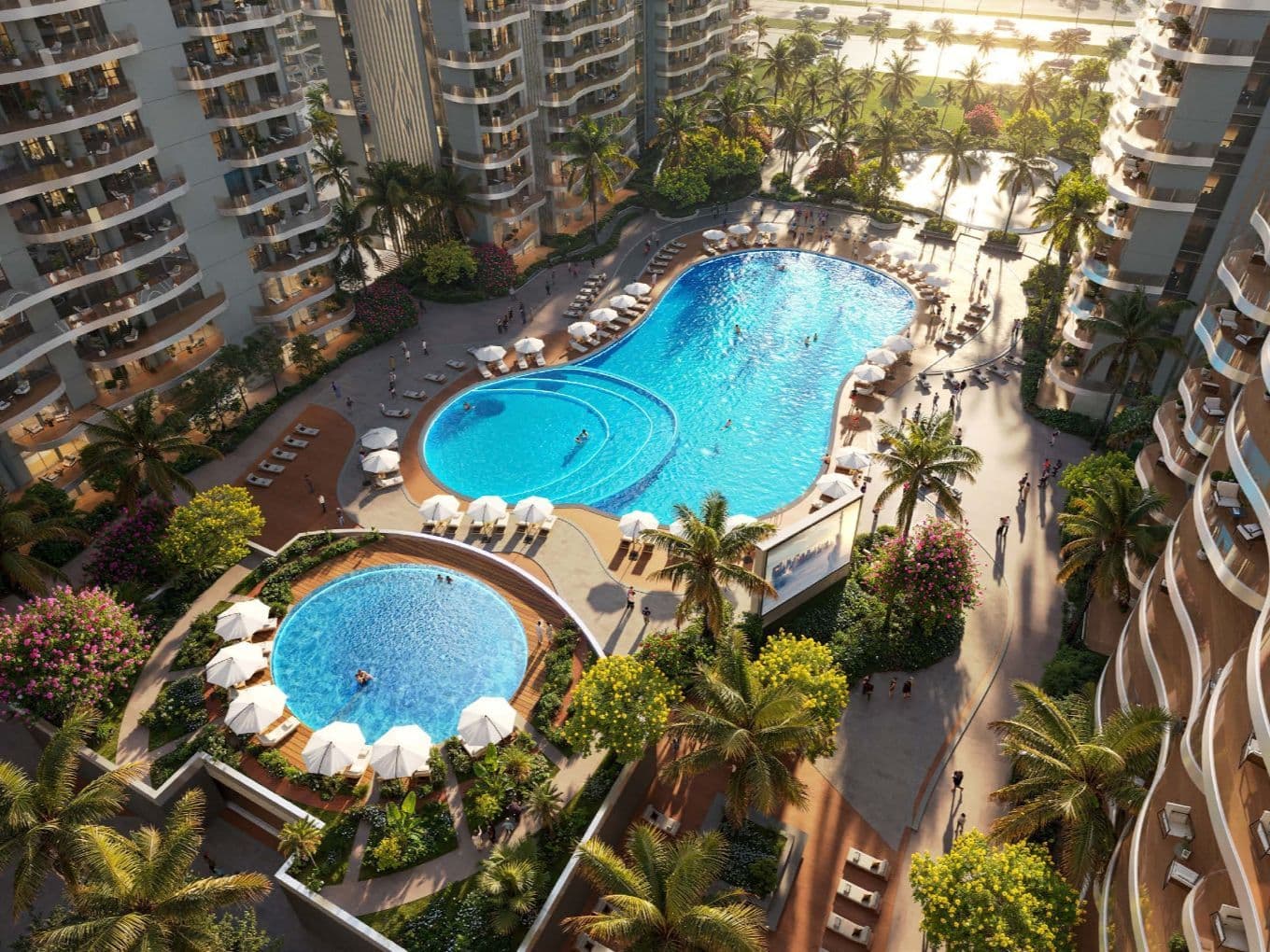 Aerial view of a modern apartment complex with two outdoor pools, surrounded by lounge chairs, umbrellas, palm trees, and gardens.
