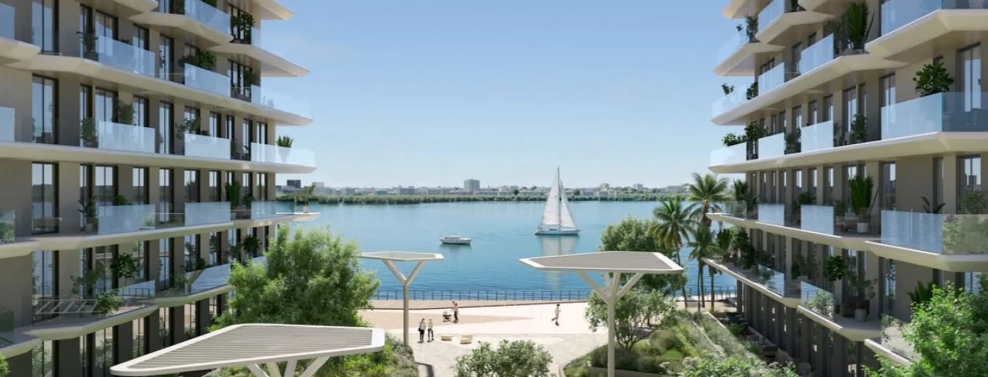 Modern waterfront apartment buildings flanking a landscaped promenade with people, palm trees and a sailboat on calm blue water.