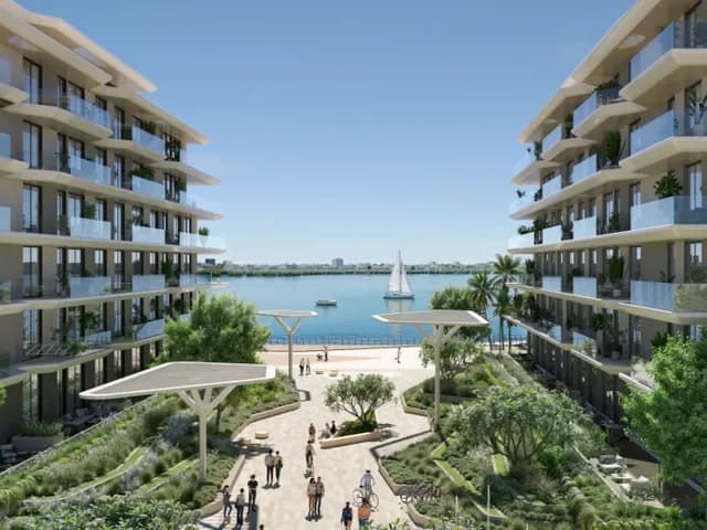 Modern waterfront apartment buildings flanking a landscaped promenade with people, palm trees and a sailboat on calm blue water.
