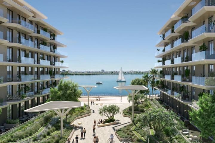 Modern waterfront apartment buildings flanking a landscaped promenade with people, palm trees and a sailboat on calm blue water.