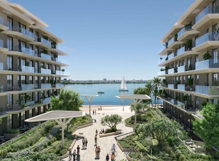 Modern waterfront apartment buildings flanking a landscaped promenade with people, palm trees and a sailboat on calm blue water.