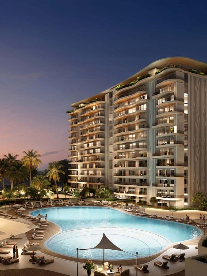 Luxurious residential complex at dusk with illuminated high-rise buildings, a large pool, palm trees, and people relaxing by lounge chairs.