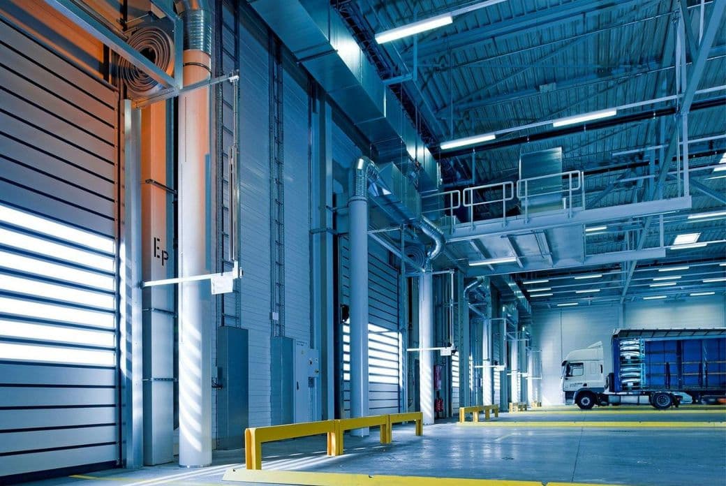 Spacious warehouse interior with high ceilings, industrial lighting, and a parked white truck near large bay doors.