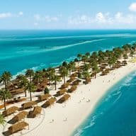 A scenic beach with palm trees, thatched-roof huts, and clear turquoise waters lining the shore under a bright blue sky.
