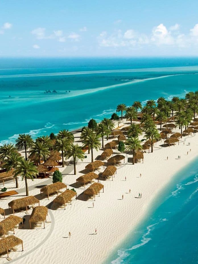 A scenic beach with palm trees, thatched-roof huts, and clear turquoise waters lining the shore under a bright blue sky.