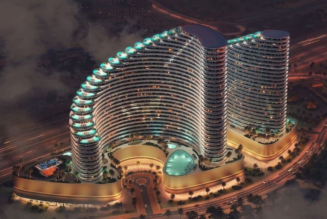 Aerial view of a modern, illuminated twin-tower complex with curved architecture, circular central pool, and surrounding roads at dusk.