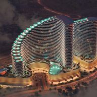 Aerial view of a modern, illuminated twin-tower complex with curved architecture, circular central pool, and surrounding roads at dusk.