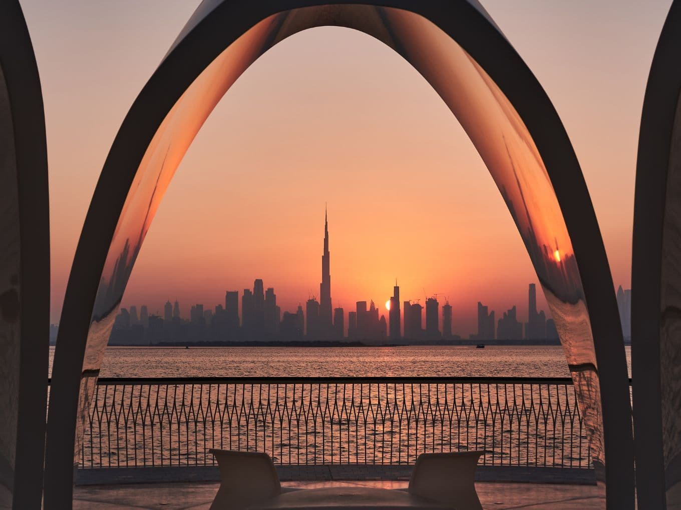 Sunset over Downtown Dubai framed by large curved arches, with Burj Khalifa, calm water, railings, and chairs in the foreground.