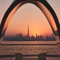 Sunset over Downtown Dubai framed by large curved arches, with Burj Khalifa, calm water, railings, and chairs in the foreground.