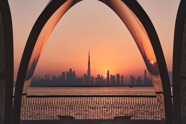 Sunset over Downtown Dubai framed by large curved arches, with Burj Khalifa, calm water, railings, and chairs in the foreground.