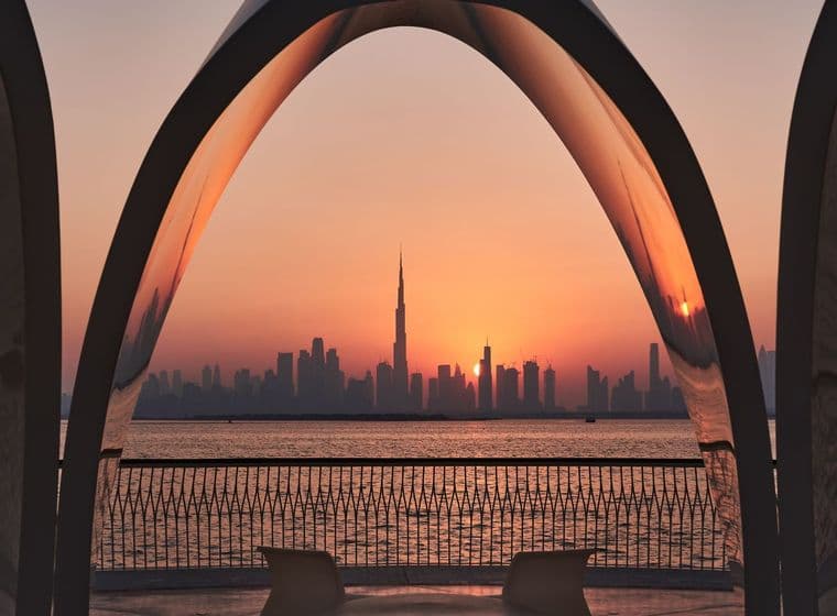 Sunset over Downtown Dubai framed by large curved arches, with Burj Khalifa, calm water, railings, and chairs in the foreground.