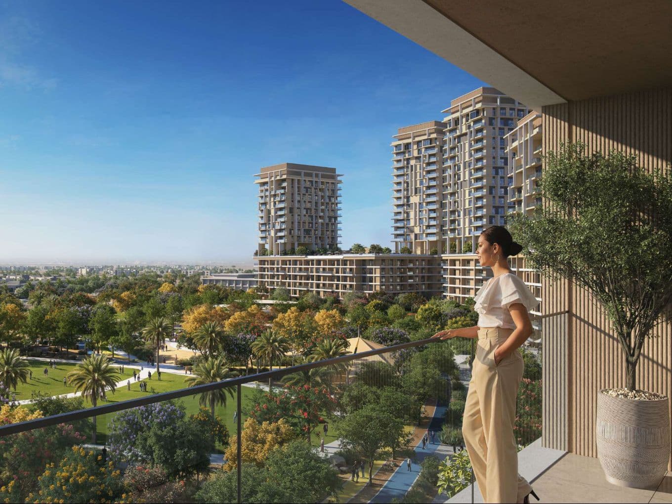 Woman on a balcony overlooking a lush park and modern high-rise apartment towers under a clear blue sky.