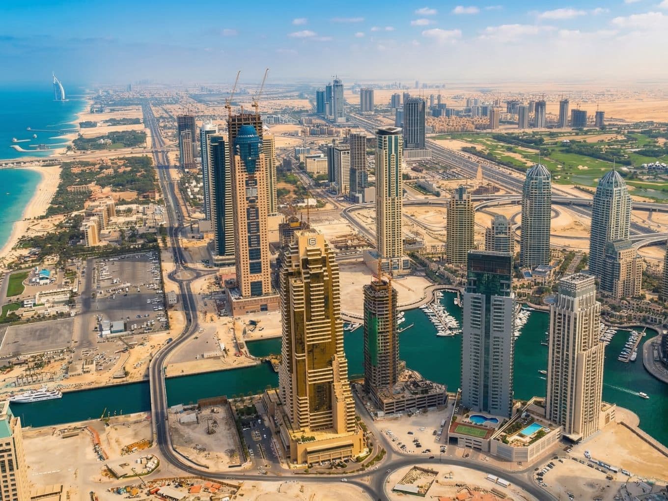Aerial view of early Dubai Marina with skyscrapers under construction, winding roads, and a canal, set against a backdrop of sand and the distant coastline.