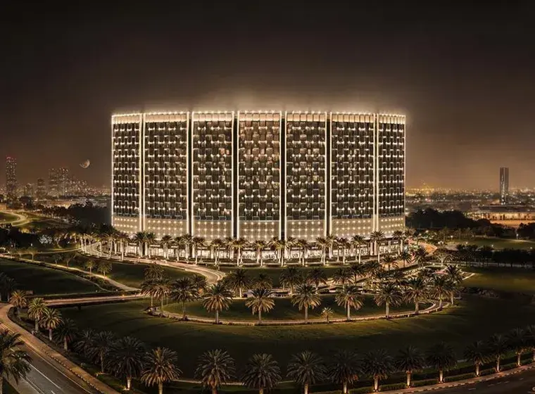 Night view of a large, illuminated building with grid-patterned windows, encircled by palm-lined roads; distant city lights in the background.