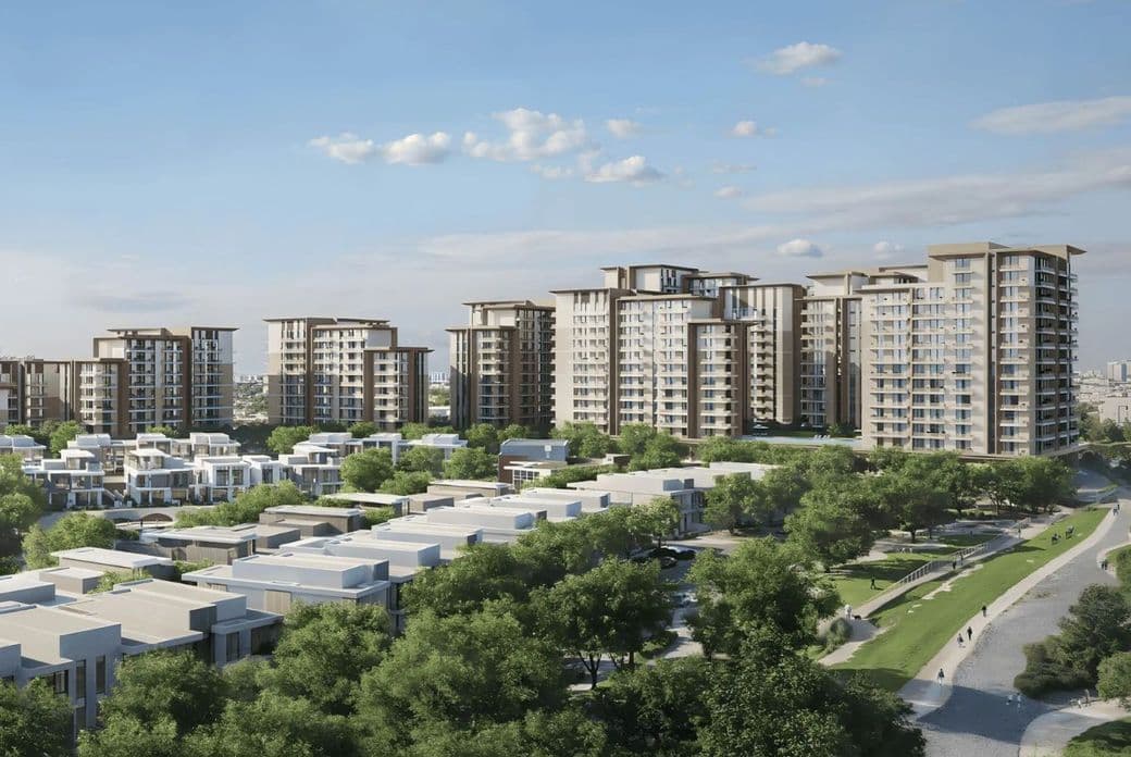 Modern apartment buildings surrounded by green trees and smaller residential structures under a blue sky with scattered clouds.
