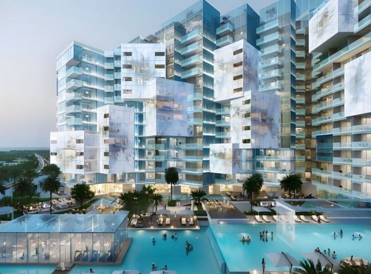 Modern, futuristic residential complex with irregularly stacked glass buildings, surrounded by pools and palm trees at dusk.