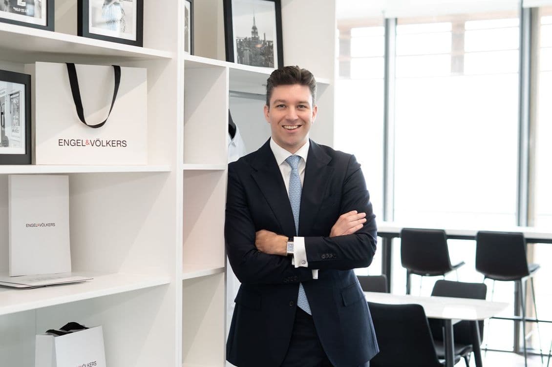 Smiling man in a dark suit and light blue tie stands with arms crossed beside white shelves displaying Engel & Völkers bags in a bright office.