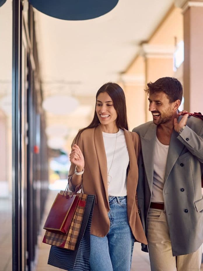 A smiling couple walks with shopping bags in an outdoor mall, dressed in casual yet stylish clothing, enjoying their day.