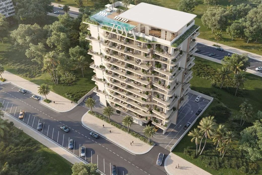 Aerial view of a modern, multi-story apartment building surrounded by trees, roads, and cars. The rooftop features greenery and a pool.