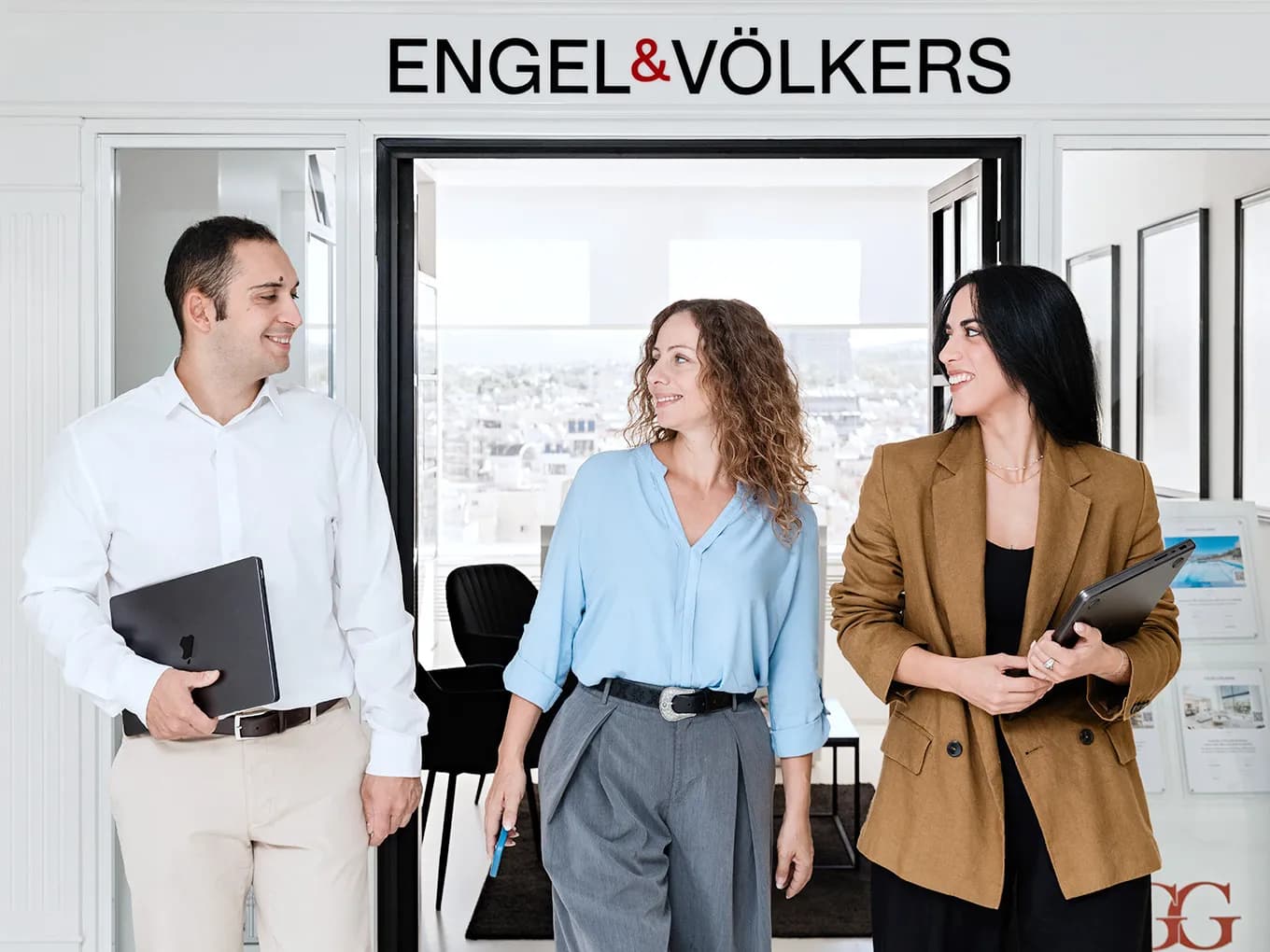 Three professionals stand in a bright Engel & Völkers office, smiling; a man holds a laptop, two women carry tablets and documents, with a city view behind.