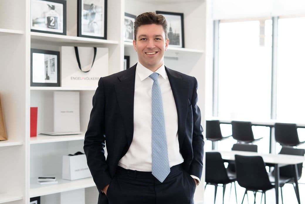 Smiling man in a dark suit and light blue tie stands with hands in his pockets in a bright office, framed photos on white shelves behind him.