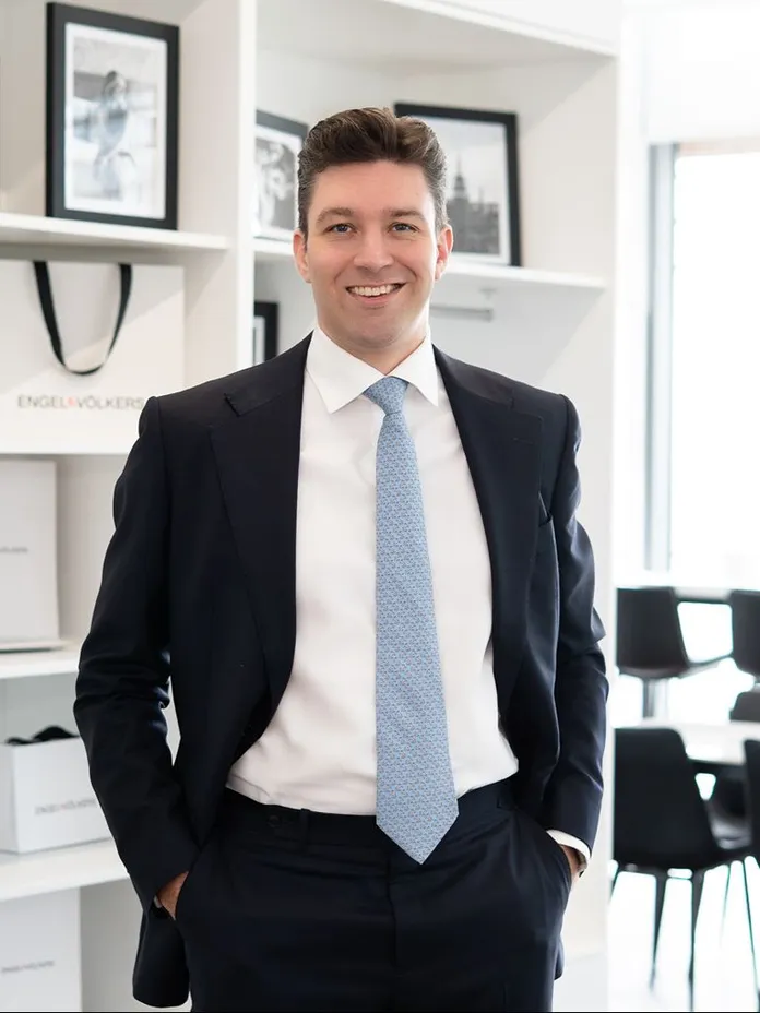 Smiling man in a dark suit and light blue tie stands with hands in his pockets in a bright office, framed photos on white shelves behind him.