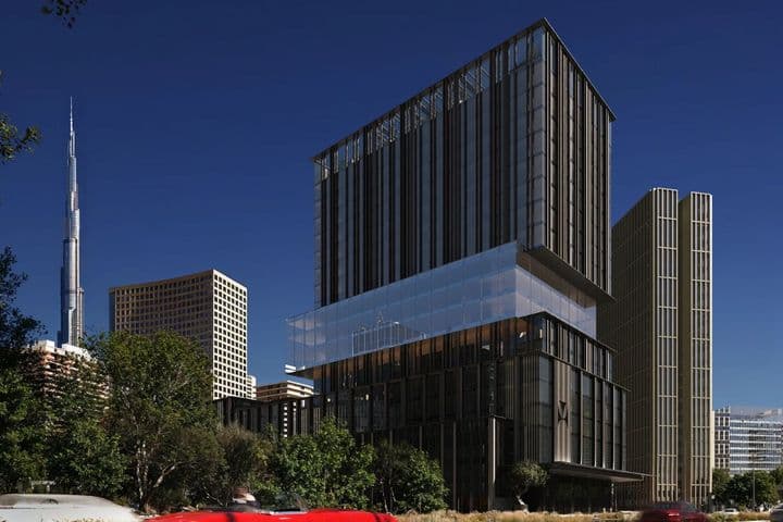Modern buildings against a clear blue sky, with a red car driving by and a tall skyscraper in the background.