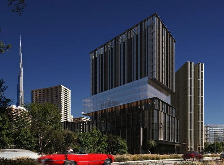 Modern buildings against a clear blue sky, with a red car driving by and a tall skyscraper in the background.