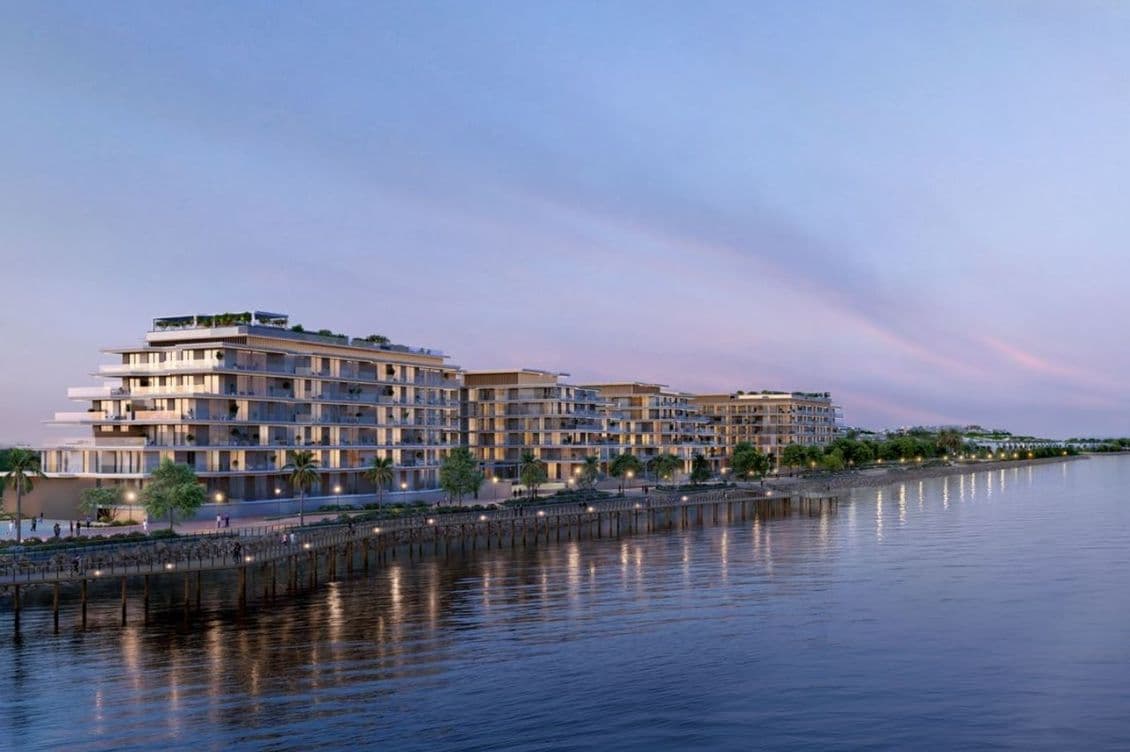 Low-rise modern waterfront apartment complex at dusk, lit balconies and a palm-lined promenade reflected on calm water.