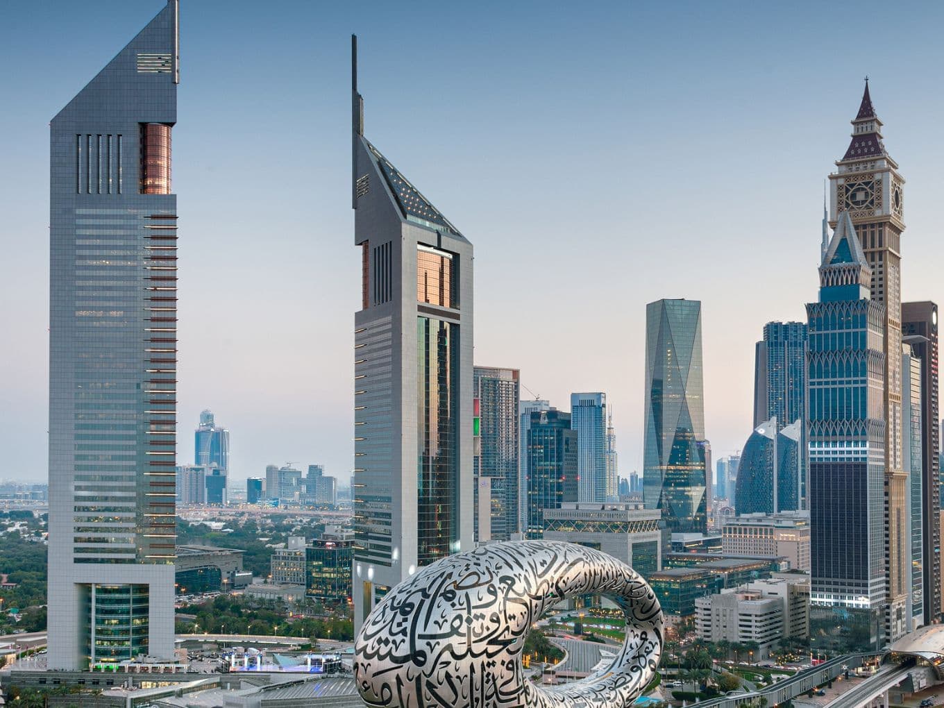 Dubai skyline with futuristic glass skyscrapers; foreground features a large white ring covered in Arabic calligraphy.