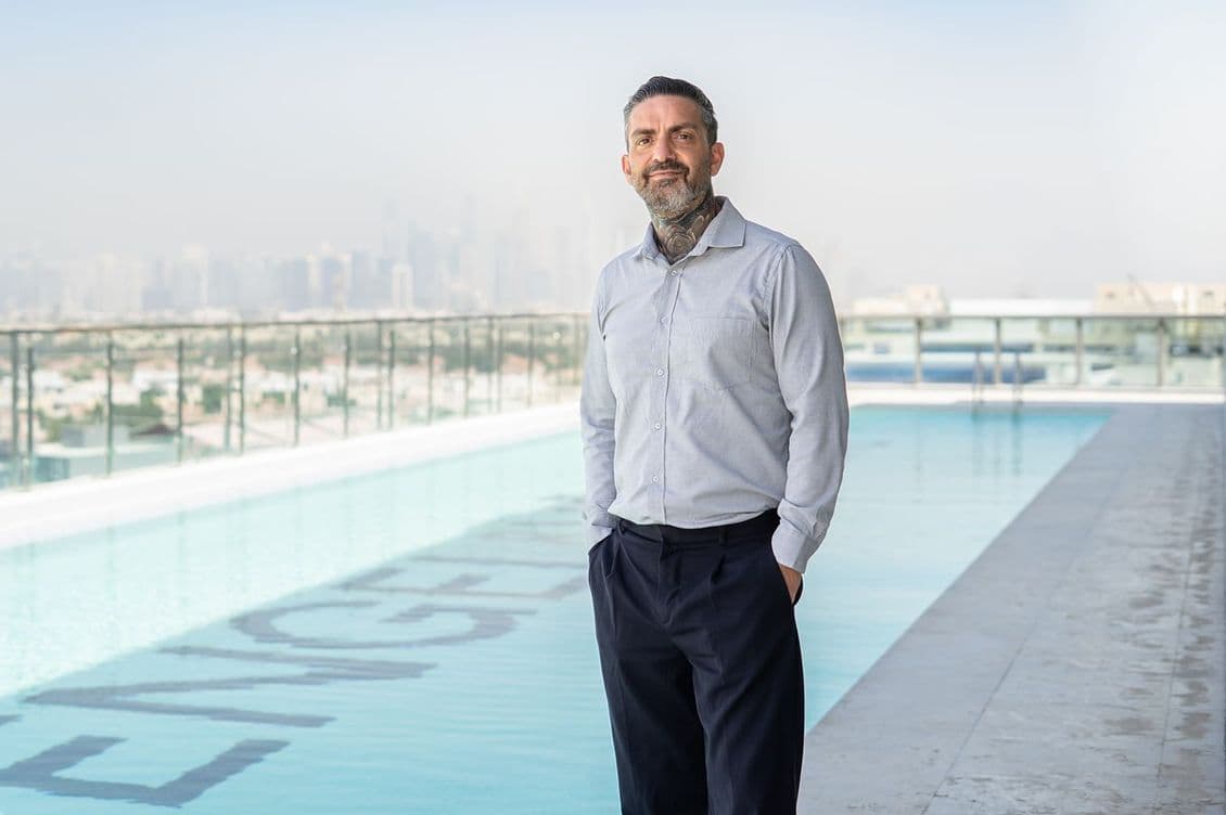 A mature man with a trimmed beard and neck tattoos stands beside a rooftop pool, wearing a light gray shirt and dark trousers, with a hazy city skyline in the background.
