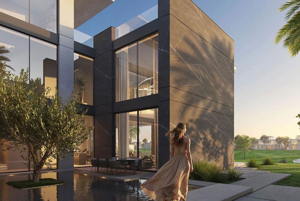 Woman in a flowing dress walks by a modern house with large windows, beside a reflective pool at sunset.