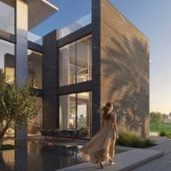 Woman in a flowing dress walks by a modern house with large windows, beside a reflective pool at sunset.