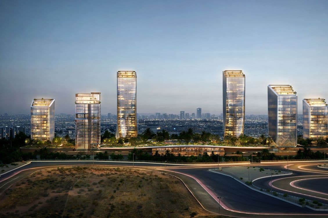 Dusk cityscape showing illuminated modern glass towers with rooftop gardens along a landscaped podium, curved highway in foreground.