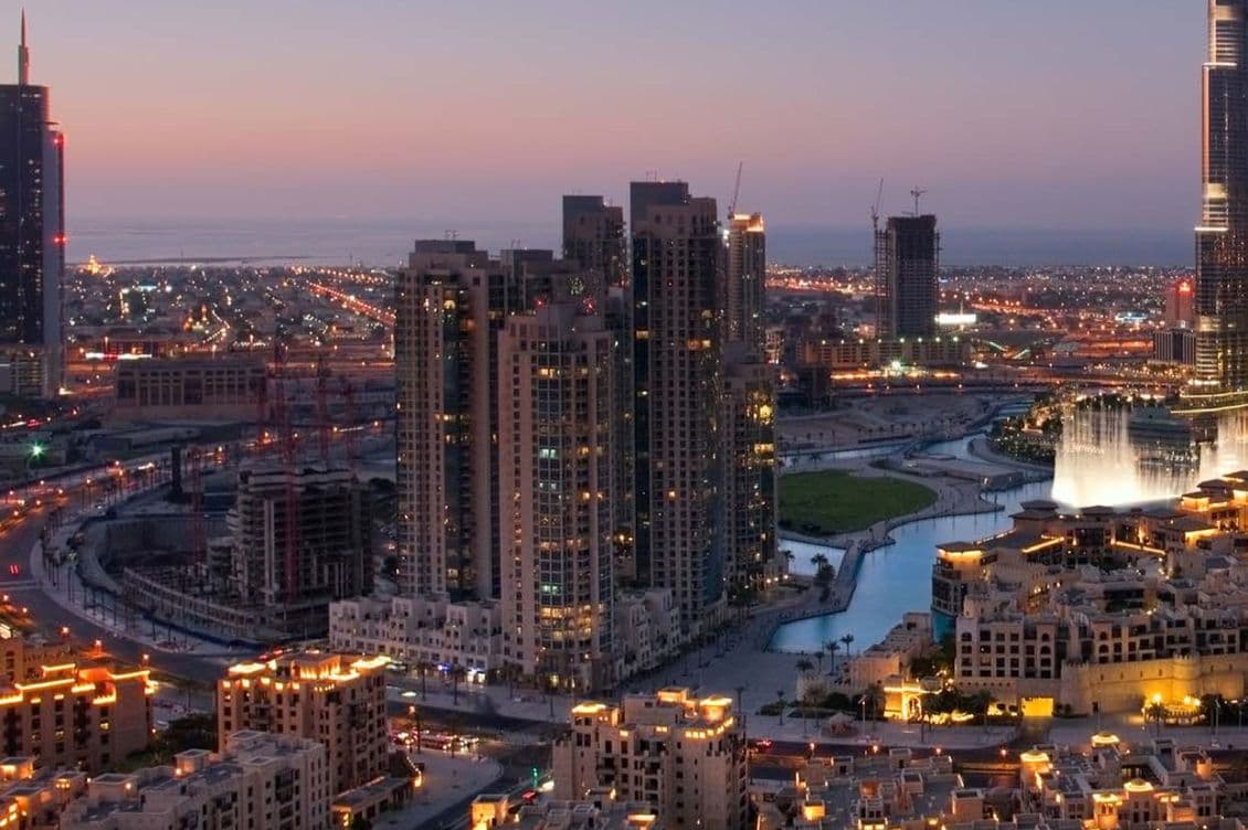City skyline at sunset with illuminated buildings, a winding waterway, and a fountain display in the foreground.
