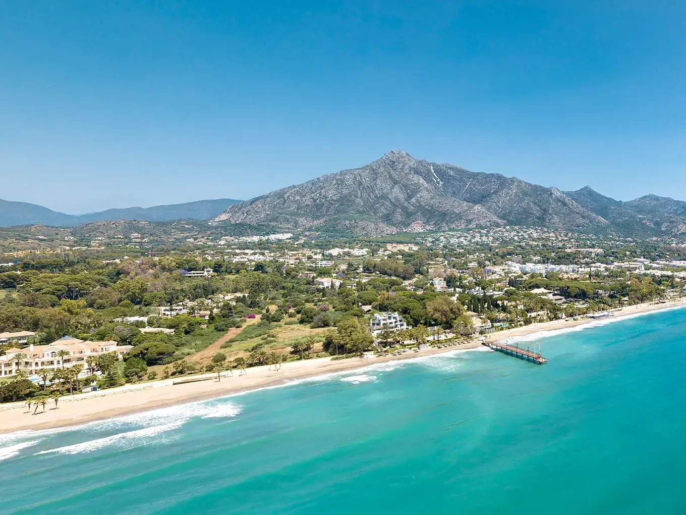 Aerial view of Marbella's coast with beach, turquoise sea, luxury villas, pier and mountains under clear blue sky
