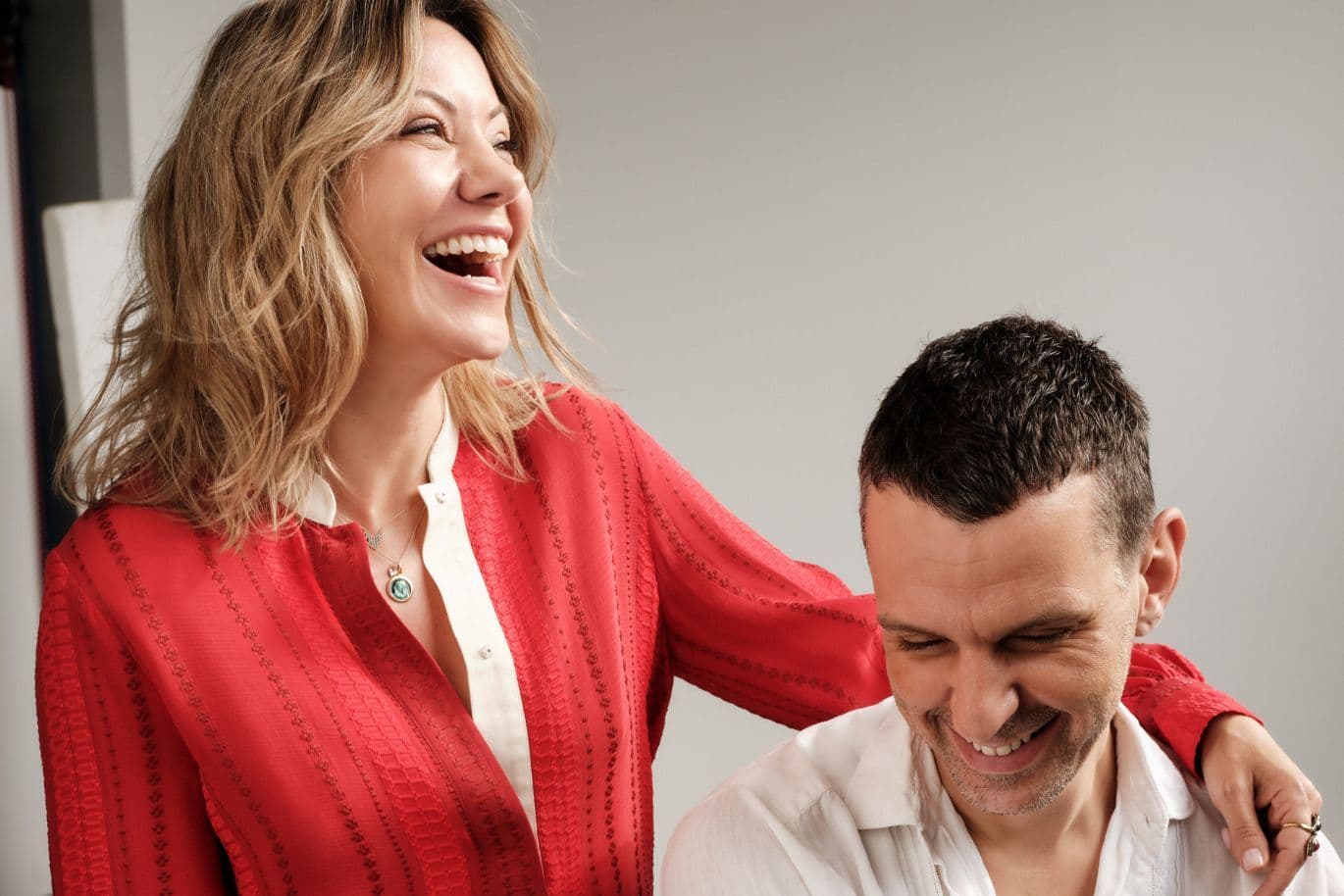 A woman in a red blouse is laughing with a man in a white shirt, who is smiling; both look happy in a brightly lit room.