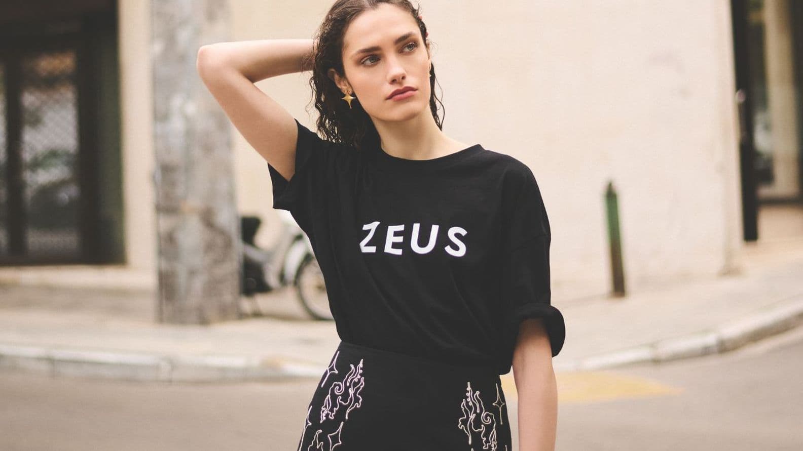 A person is standing on the street, wearing a black “ZEUS” T-shirt and a patterned skirt, with one hand on their head.