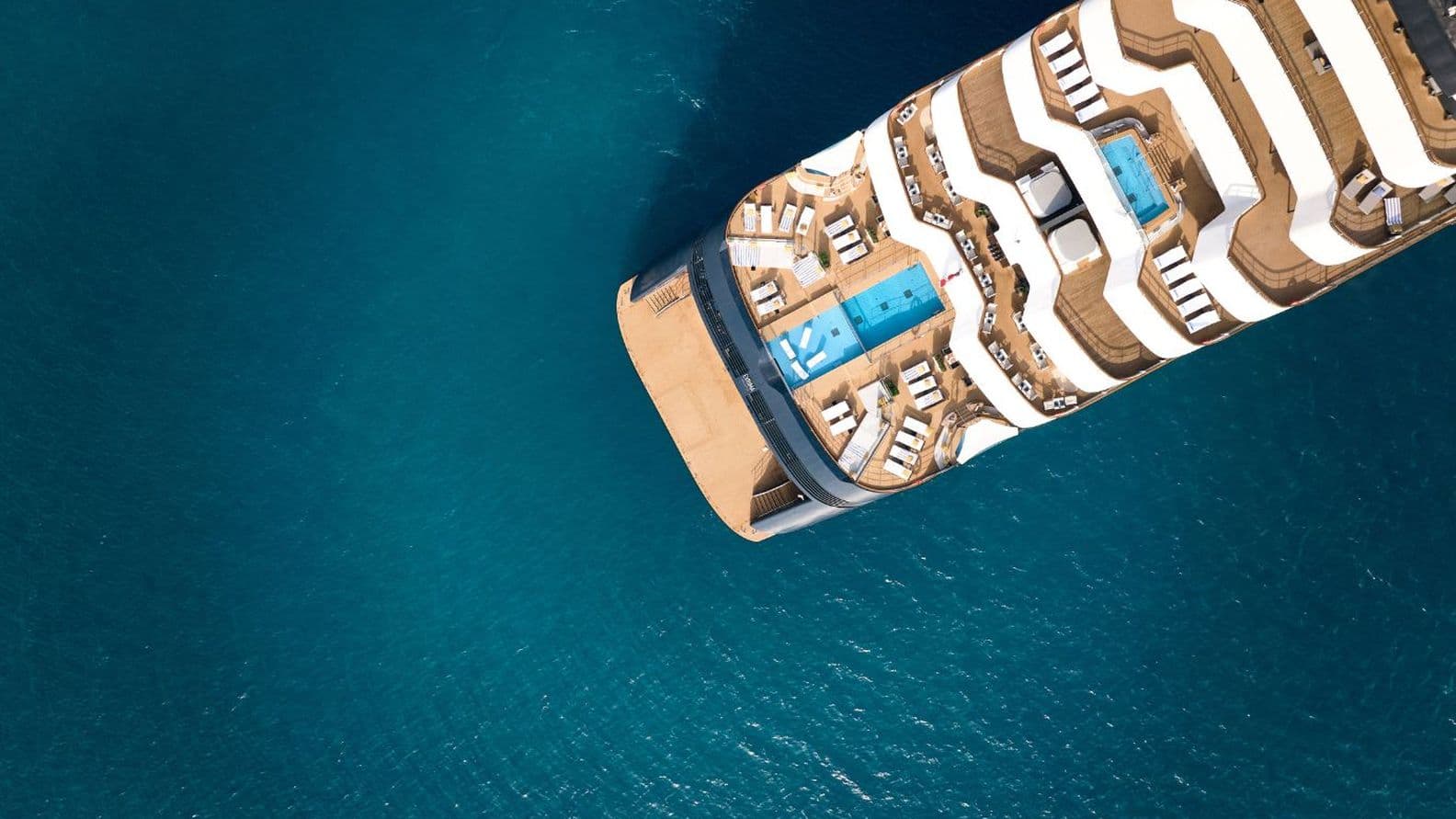 Luftaufnahme des Sonnendecks eines Kreuzfahrtschiffes mit Pools und weißen Liegestühlen auf hellbraunem Deck, umgeben vom tiefblauen Meer.