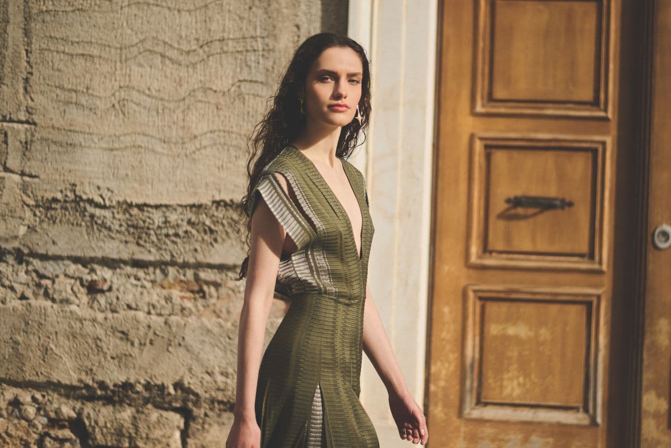 A woman in a green textured dress walks outside alongside a wooden door and a textured wall, while the sunlight casts soft shadows.