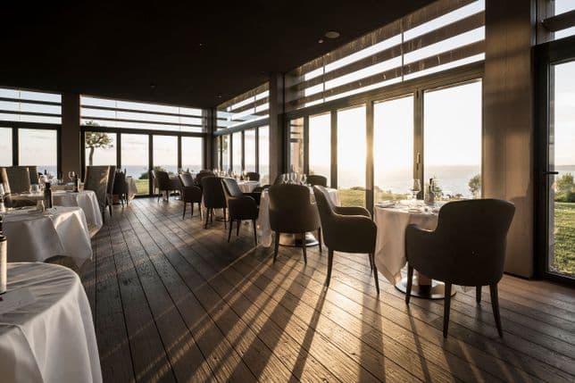 Sonnenüberflutetes Interieur eines Restaurants am Meer mit weiß gedeckten Tischen, gemütlichen Sesseln und raumhohen Fenstern mit Blick auf den Ozean bei Sonnenuntergang